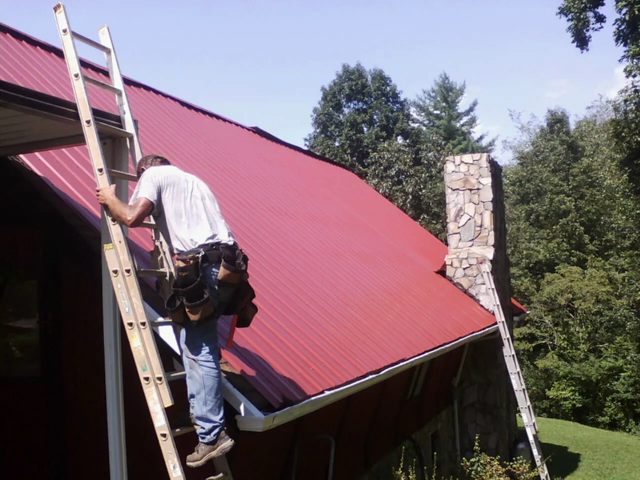 Roofer on a ladder beside a red metal roof and stone chimney — Soffit & Fascia Repair by Roofing Brothers in Royal City